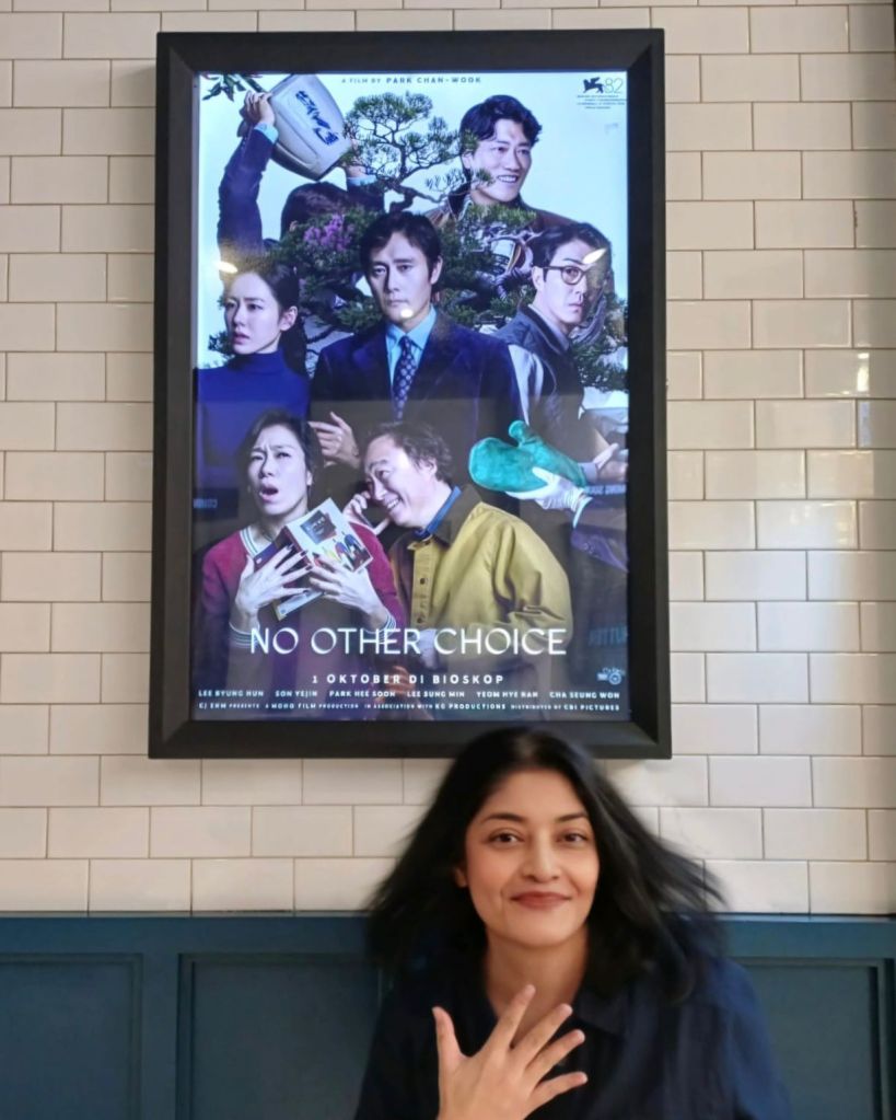 A woman smiling in front of a movie poster for the film 'No Other Choice' directed by Park Chan-wook, featuring multiple characters in a collage style.