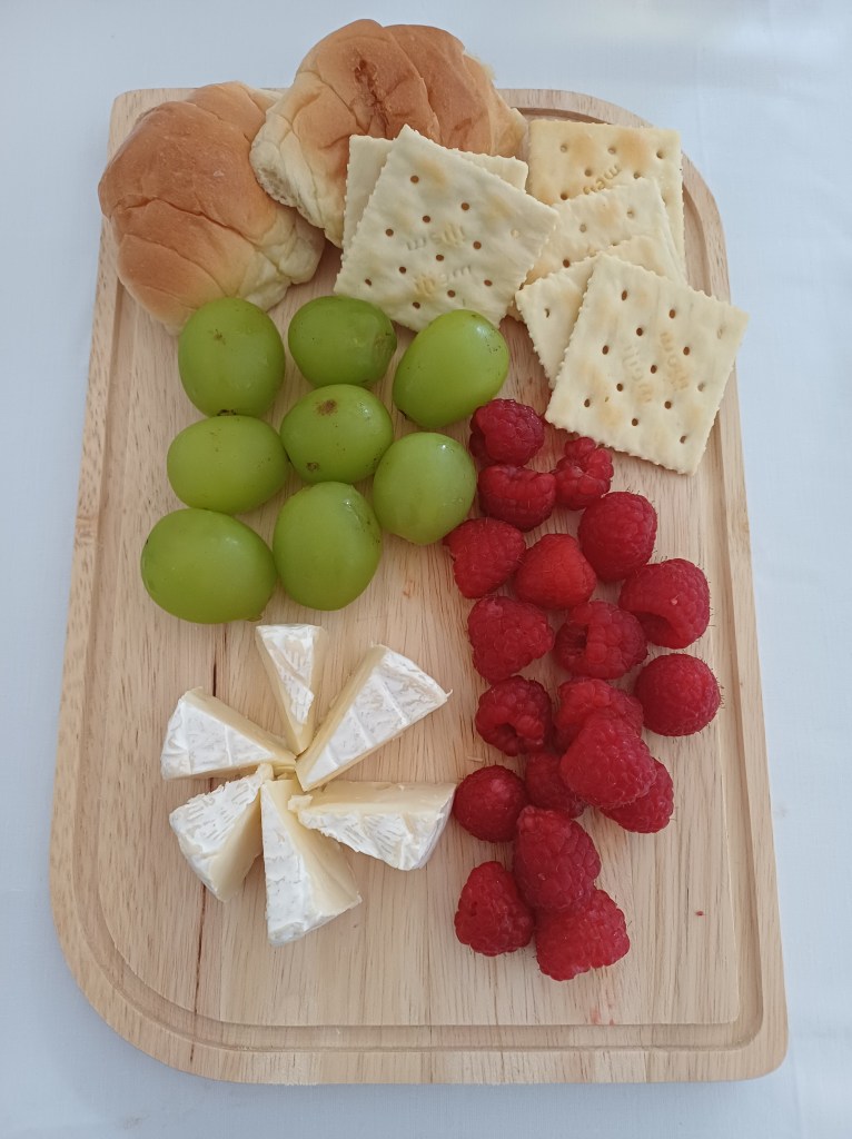 A wooden platter featuring a selection of homemade cheese, green grapes, raspberries, crackers, and soft bread rolls.